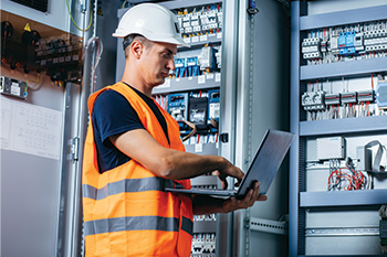 Technician using a laptop to inspect an electrical control panel.