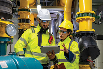 Technicians inspecting chiller equipment with tablet among industrial pipes and gauges