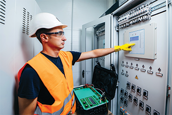 Electrical technician wearing safety gear inspecting and testing an industrial control panel in a manufacturing facility.